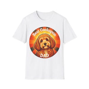 Puede incluir: Una camiseta blanca de manga corta con un gráfico circular en el pecho. El gráfico muestra un perro Cockapoo marrón centrado sobre un fondo de rayos de sol rojos y naranjas, con el texto blanco Best Cockapoo DAD arqueado alrededor.