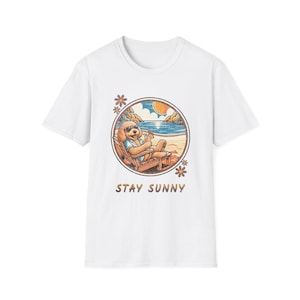 Peut inclure: Un t-shirt blanc à manches courtes avec un motif circulaire représentant un chien doré portant des lunettes de soleil, se relaxant dans une chaise de plage avec une boisson tropicale. Le texte STAY SUNNY est imprimé sous le motif.
