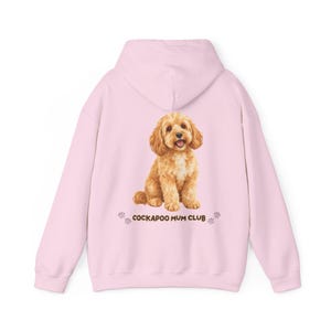 Pode incluir: Um moletom com capuz rosa claro apresentando uma ilustração realista de um cão Cockapoo castanho-dourado sentado ao centro. Abaixo do cão, o texto COCKAPOO MUM CLUB em letras maiúsculas castanhas, ladeado por pequenos ícones de patas.