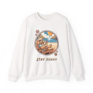 Könnte beinhalten: Ein weißes Langarm-Sweatshirt mit einer kreisförmigen Grafik eines goldenen Hundes mit Sonnenbrille, der in einem Strandstuhl mit einem Getränk entspannt. Unter der Grafik befindet sich der Schriftzug STAY SUNNY.