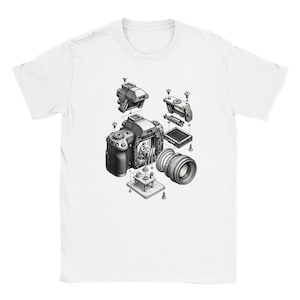 May include: A white short-sleeve t-shirt featuring a black and white graphic print of a disassembled camera. The illustration shows the camera body, lens, and internal components separated, with a tiny person painting inside the camera chamber.
