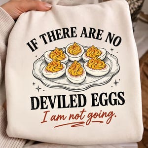 May include: A cream-colored sweatshirt featuring a graphic of five deviled eggs on a white plate. The text reads IF THERE ARE NO DEVILED EGGS I am not going. The design uses black and red lettering with a detailed illustration of the food.