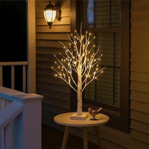 May include: A white birch-style decorative tree lamp featuring numerous small, warm-glowing LED lights on its branches. The tree stands on a square base and is accompanied by a small, metallic bird figurine placed on the circular tabletop surface.