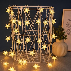 Puede incluir: Una cadena de luces LED con múltiples cubiertas translúcidas en forma de estrella de cinco puntas. Las luces emiten un brillo dorado cálido y están dispuestas sobre una estructura de cesta de alambre blanco y geométrica.