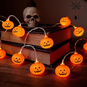 Peut inclure: Une guirlande lumineuse composée de petites citrouilles orange avec des yeux triangulaires et des bouches dentelées sculptées. Chaque citrouille est éclairée de l'intérieur et reliée par un fil transparent fin pour une décoration.