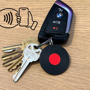 May include: A black BMW car key fob with a set of metal keys attached to a metal ring. A circular black keychain with a red center resembling a vinyl record is attached to the ring. A black graphic icon of a hand holding a phone with signal waves is visible.