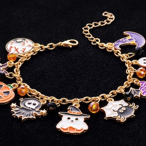 Peut inclure: Un bracelet à breloques en chaîne dorée avec des pendentifs en émail représentant une citrouille, un fantôme, une araignée sur une toile, une chauve-souris, un cupcake et un chapeau de sorcière, agrémenté de perles orange et noires.