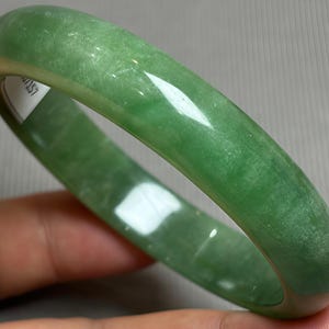 Pode incluir: Uma pulseira rígida verde, lisa e polida, com um perfil arredondado. A superfície da pulseira apresenta variações naturais de cor, variando do verde esmeralda claro ao profundo, com um acabamento brilhante que reflete a luz na sua superfície.