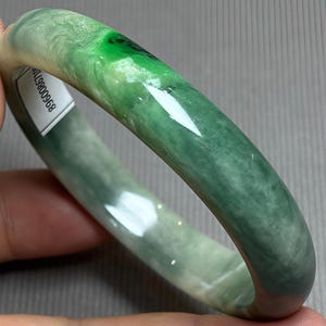 Pode incluir: Uma pulseira de jade lisa e polida com um padrão variado de tons verde-floresta profundo e verde-claro translúcido. Uma pequena etiqueta retangular branca com o número 896008968 impresso em preto está fixada na superfície interna.