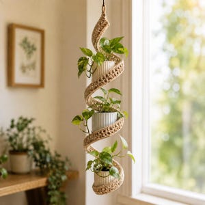 May include: A hanging plant holder featuring a spiral design made of tan woven rope. The structure holds three white, vertically ribbed cylindrical pots, each containing a green pothos plant with heart-shaped leaves, suspended vertically.