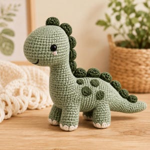 Puede incluir: Un juguete de peluche de dinosaurio tejido a ganchillo en color verde salvia con cuello largo, una hilera de espinas redondeadas de color verde oscuro en la espalda, manchas circulares verde oscuro en el costado, ojos de cuentas negras y detalles blancos en pies y mejillas.