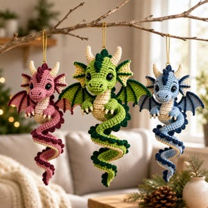 May include: Three crocheted dragon ornaments hanging from a branch. The left dragon is pink with dark pink accents, the centre dragon is green with dark green accents, and the right dragon is light blue with dark blue accents. Each has horns.