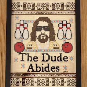 May include: A framed cross-stitch design featuring a cartoon image of The Dude from the film The Big Lebowski. The Dude is wearing sunglasses and a white shirt. The text "The Dude Abides" is below the image.