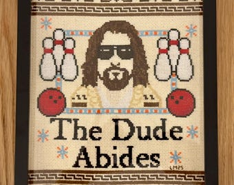 May include: A framed cross-stitch design featuring a cartoon image of The Dude from the movie The Big Lebowski. The Dude is wearing sunglasses and a white shirt. The text "The Dude Abides" is below the image.