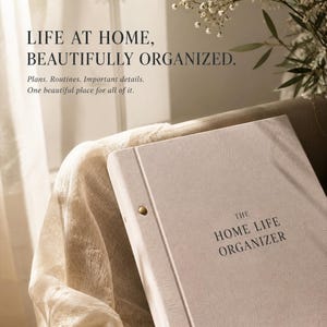 Puede incluir: Una carpeta cubierta de tela beige claro titulada THE HOME LIFE ORGANIZER con remaches metálicos dorados a lo largo del lomo. El texto LIFE AT HOME, BEAUTIFULLY ORGANIZED. Plans. Routines. Important details. One beautiful place for all of it. es visible.