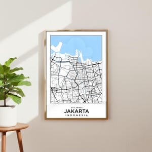 Op de afbeelding: Een ingelijste wandkunstprint met een minimalistische kaart van Jakarta, Indonesië. Het ontwerp toont zwarte straatlijnen op een witte achtergrond met een lichtblauw watergedeelte aan de bovenkant. De tekst luidt 6°7' S, 106°46' E, JAKARTA, INDONESIA.