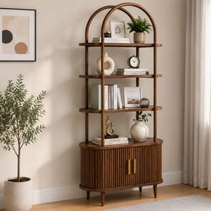 May include: A tall, arched wooden shelving unit featuring four open shelves and a lower cabinet with two fluted doors and gold handles. The shelves are decorated with various books, a white ceramic vase, a clock, and small potted plants.