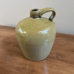 May include: A ceramic jug with a rounded body and a single handle. The upper portion features a glossy, mottled olive-green glaze, while the lower section transitions into a matte, light beige finish. A dark, textured band encircles the neck.