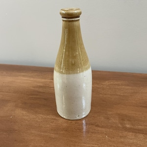 May include: A ceramic bottle with a cylindrical body and a narrow neck. The upper portion of the bottle features a tan-colored glaze, while the lower portion is finished in an off-white glaze. The bottle has a rounded rim at the top opening.
