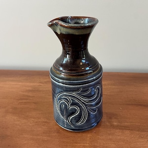May include: A ceramic pitcher with a dark brown glazed neck and a blue-grey glazed body. The cylindrical base features a raised, embossed swirling heart pattern. The vessel has a flared rim with a small pouring spout and a smooth, glossy finish.