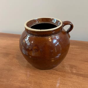 May include: A round, brown glazed ceramic pot with a single side handle and a wide, open top. The exterior features a glossy, dark brown finish with subtle variations in tone, while the inner rim is a contrasting light cream color.