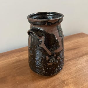 May include: A cylindrical ceramic vessel with a wide, flared rim and a small handle on one side. The exterior features a contrasting design of dark, glossy black glaze applied over a matte, earthy brown clay base, creating a textured appearance.