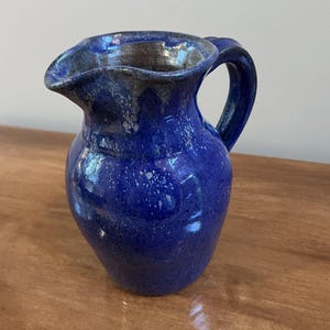 May include: A ceramic pitcher with a glossy, deep blue glaze and a curved handle. The rim features a textured, mottled grey and blue finish that drips down the neck. The body of the pitcher is rounded and smooth, showcasing a vibrant cobalt blue color throughout the exterior surface.