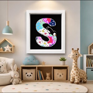 May include: A square white-framed wall art piece featuring a large, stylized letter S on a black background. The letter is decorated with colorful patterns, including blue, pink, and white sections, and features a small black and white penguin illustration.