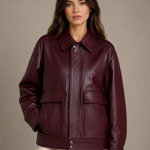 May include: A deep burgundy leather jacket featuring a classic collar, long sleeves, and two large front patch pockets with flaps. The jacket includes a front placket with visible silver-toned snap button closures at the hem and collar.