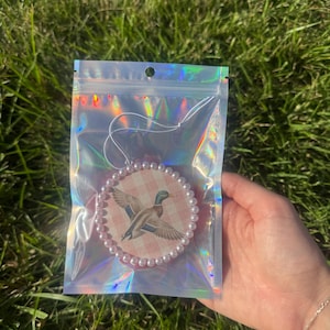 Puede incluir: Un adorno colgante circular con una ilustración de un pato azulón sobre un fondo de cuadros vichy rosa y blanco, bordeado por un anillo de pequeñas perlas blancas, dentro de una bolsa de almacenamiento holográfica transparente con cordón blanco.