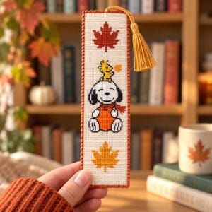 May include: A rectangular cross-stitch bookmark on white fabric with a red maple leaf at the top, a yellow maple leaf at the bottom, and a central design of Snoopy wearing an orange sweater with Woodstock on their head, finished with a yellow tassel.