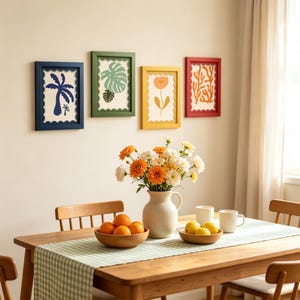 May include: Four rectangular wall frames with scalloped inner edges in blue, green, yellow, and red, each containing a botanical print. A white ceramic pitcher filled with orange and white flowers sits on a wooden table with two bowls of oranges.