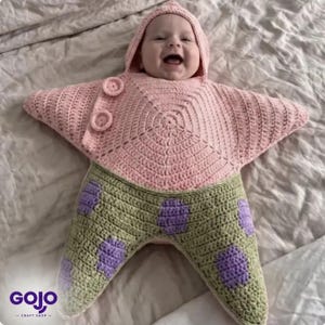 May include: A crochet baby bunting shaped like a starfish with a hood. The top section is light pink with two circular buttons, and the bottom section is light green with purple oval spots. The garment features a star-like silhouette.