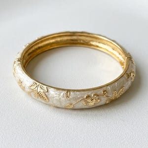 Op de afbeelding: Een scharnierende armband met een basis van gepolijst goudkleurig metaal en een crème-kleurige emaille inleg. Het oppervlak is versierd met reliëf goudkleurige bloem- en bladmotieven die rond de buitenkant van de cirkelvormige band lopen.