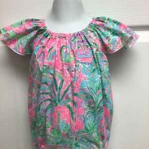 May include: A colorful romper with a tropical print featuring pink buildings, palm trees, and giraffes on a light blue background. The romper has short, ruffled sleeves and an elasticated neckline and waist.
