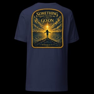 May include: A navy blue t-shirt featuring a graphic on the back. The design shows a person standing on a mountain peak with arms outstretched toward a bright light in the sky. Text reads: SOMETHING KEEPS TELLING ME TO GO ON and ROMANS 8:31.