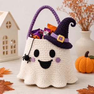 Può includere: Un cestino a forma di fantasma all'uncinetto color crema con manico viola, caratterizzato da un ragno nero appeso a un filo, due occhi neri, un ampio sorriso nero e guance rosa. Un cappello da strega viola scuro con fibbia gialla è posato.