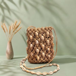 May include: A tan macrame crossbody bag featuring a knotted cord design embellished with numerous white cowrie shells throughout the body and the shoulder strap, paired with a tall, slender, textured tan ceramic vase holding dried wheat stalks.