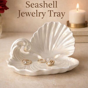May include: A white, pearlescent seashell jewelry tray featuring a sculpted wave design on one side. The tray holds two gold rings and a small gold stud earring. The text "Seashell Jewelry Tray" is printed in brown above the object.