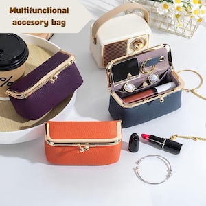 May include: Three small, rectangular, textured accessory bags with gold-tone metal kiss-lock frames in purple, orange, and dark blue. The open dark blue bag reveals a small mirror, two gold rings, and a lipstick. A cream-colored radio-shaped case is nearby.