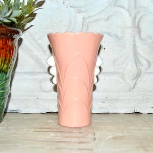 Pode incluir: Um vaso rosa claro com uma borda ondulada e detalhes brancos e arredondados. O vaso tem uma forma alargada e um padrão vertical decorativo. O vaso é exibido em uma superfície de madeira.