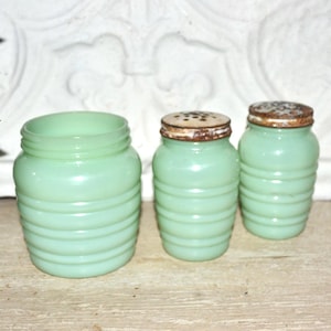 May include: A set of three vintage jadeite glass kitchen canisters. The canisters have a ribbed design and are in varying sizes. Two of the canisters have metal lids with small holes. The glass has a soft, opaque green color.