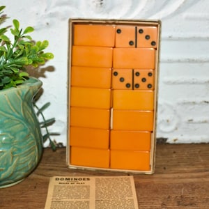 May include: A vintage set of orange dominoes in a cardboard box, with black dots. The box is open, revealing the tiles. A printed rules sheet is in front of the box. A green ceramic pot with a succulent plant is to the left.