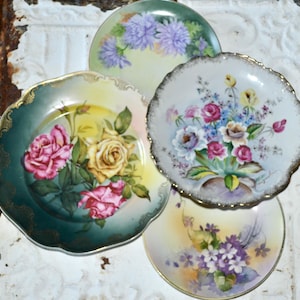 May include: A collection of four round porcelain plates featuring floral designs. One plate has pink and yellow roses on a green background, another displays a bouquet of roses, and two others feature purple and white flowers with gold trim.