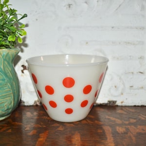 May include: White glass mixing bowl with red polka dots. The bowl has a flared shape and is likely vintage. The bowl is sitting on a wooden surface. A green vase with a plant is to the left of the bowl.