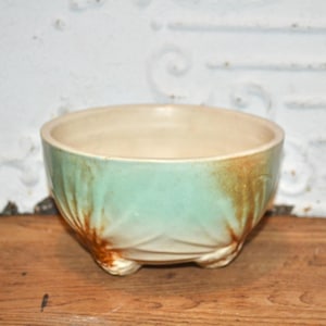 Pode incluir: Uma tigela de cerâmica com exterior verde claro e interior bege. A tigela tem um design texturizado e assenta em três pequenos pés. O exterior tem detalhes em castanho, o que contribui para a sua estética vintage.