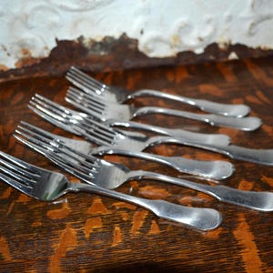 May include: A set of four silver-toned metal forks arranged in a staggered formation. Each fork features a polished finish, four tines, and a smooth, rounded handle design. The forks are positioned diagonally, showcasing their reflective surfaces.
