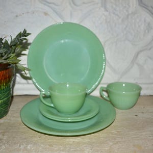 Puede incluir: Una colección de vajilla vintage de jadeíta, que incluye platos, platillos y tazas de té. La vajilla es de un color verde claro y suave. Un macetero decorativo con follaje verde está en el fondo.
