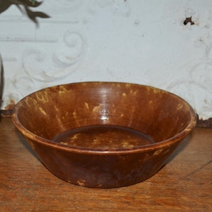 Pode incluir: Uma tigela de cerâmica marrom com uma borda larga. A tigela tem um acabamento brilhante e um padrão salpicado. A tigela está em uma superfície de madeira. A tigela é provavelmente usada para servir comida.