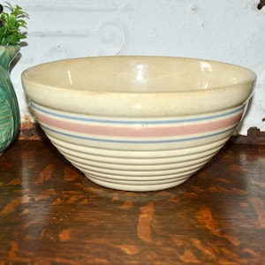May include: A cream-colored ceramic mixing bowl featuring a ribbed exterior texture. The bowl is decorated with a wide horizontal pink band flanked by two thinner blue stripes encircling the middle section of the vessel's rounded body.